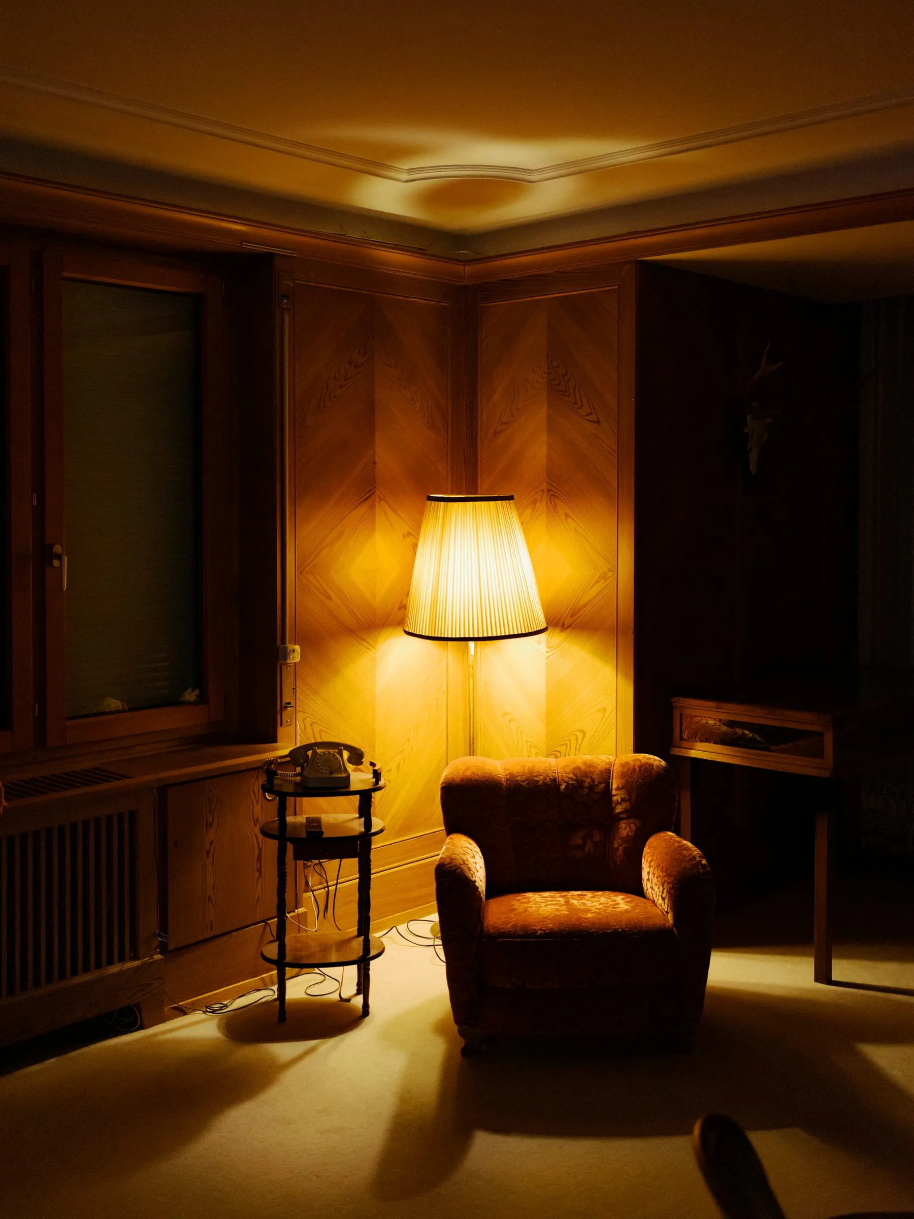 Ein holzgetäfeltes Wohnzimmer der Villa, von einer Stehlampe warm ausgeleuchtet, menschenleer.