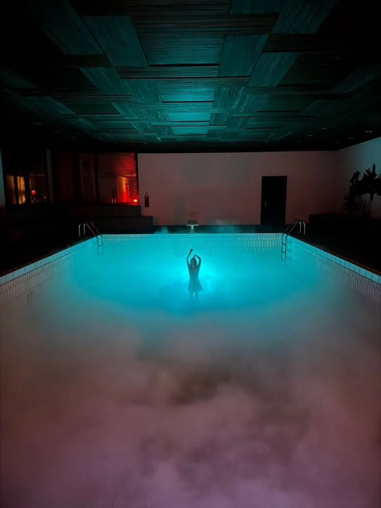 Dasselbe Hallenbad in türkisfarbenem Licht, dichter Nebel liegt über dem Becken, in der Mitte eine Figur mit erhobenen Armen.