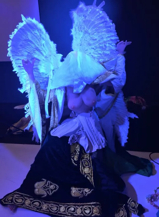 Eine Figur mit großen weißen Federflügeln kniet in blauem Licht auf einem gemusterten Teppich.
