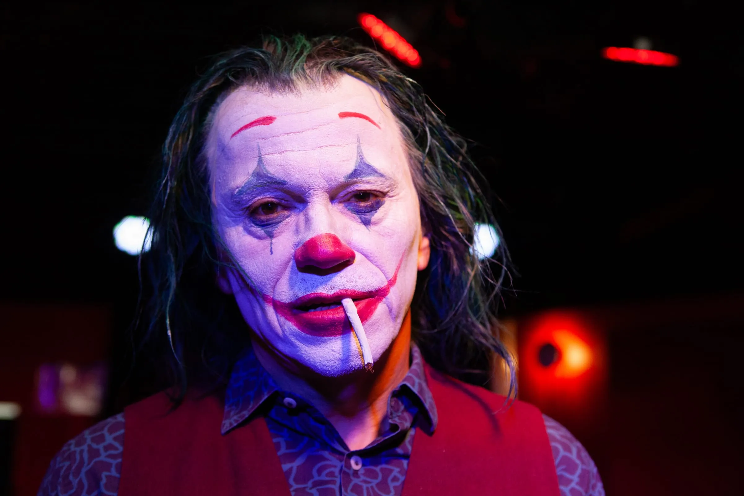 Nahaufnahme des Jokers mit weiß geschminktem Gesicht, rotem Mund und grün gefärbtem Haar im roten Bühnenlicht.
