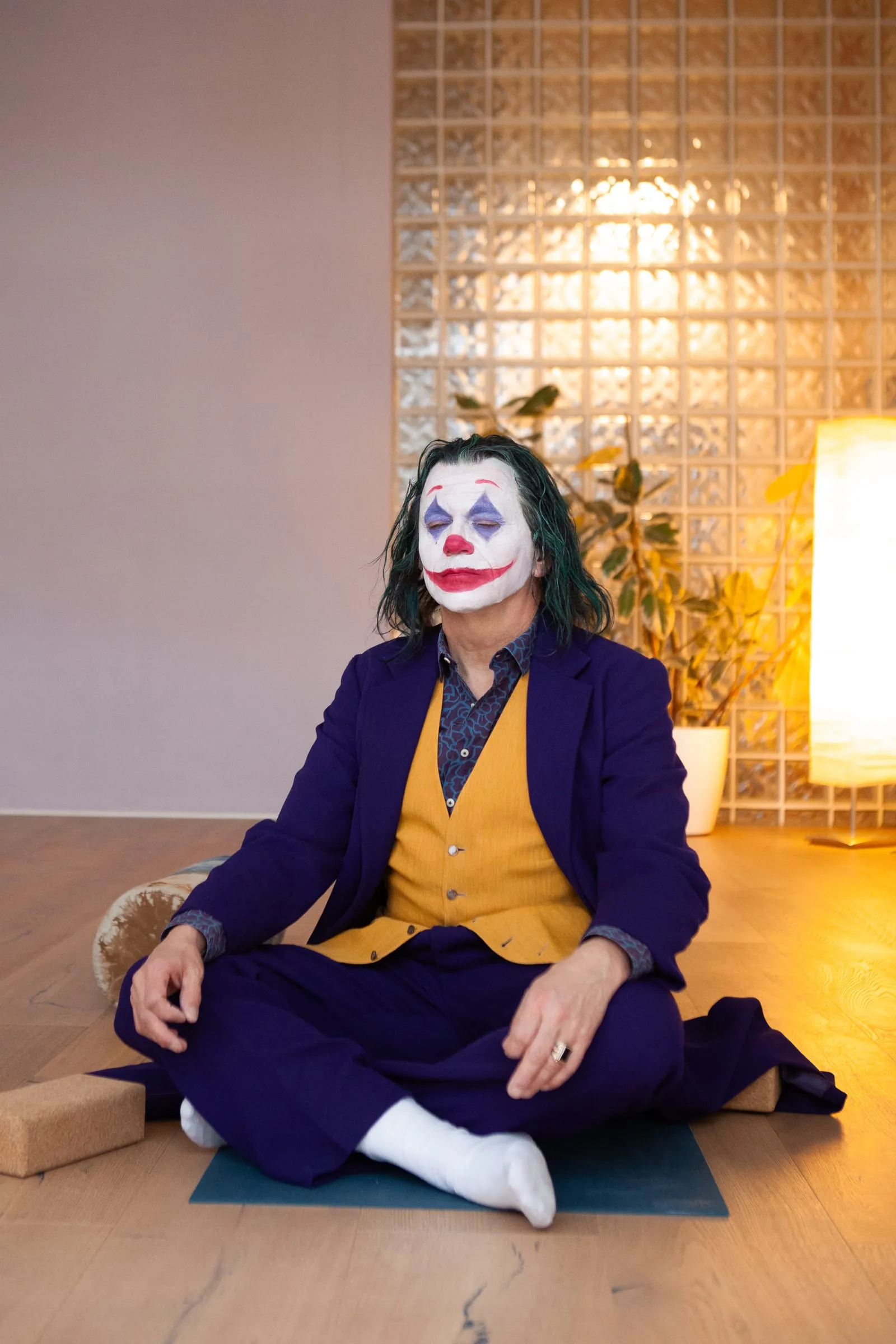 Der Joker sitzt im Schneidersitz auf einer Matte in einem hellen Raum, daneben eine Stehlampe.