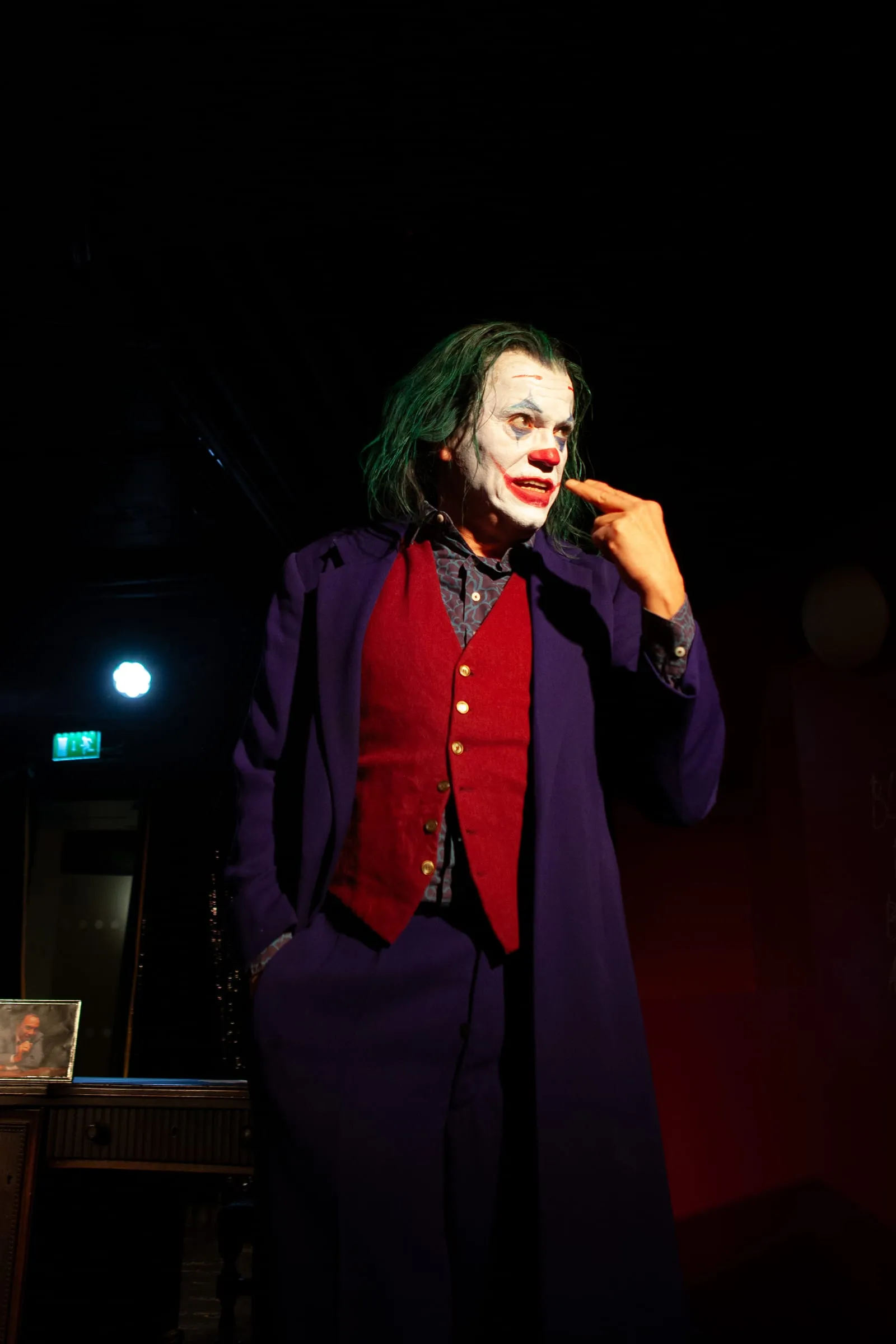 Der Joker in violettem Mantel und roter Weste fasst sich im Halbdunkel ans Gesicht.
