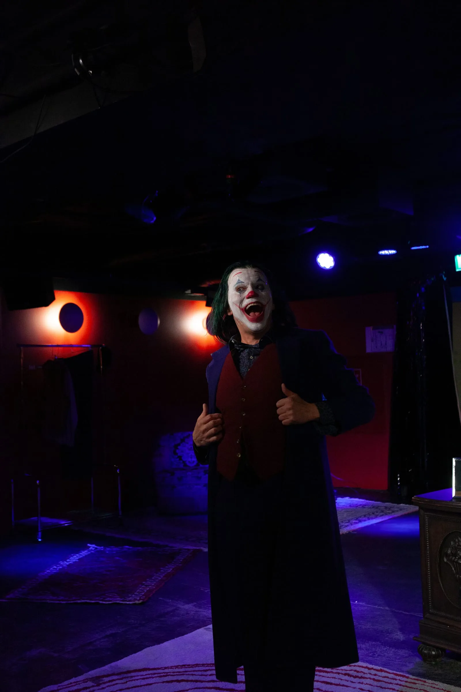 Der Joker steht lachend im blau ausgeleuchteten Barraum, den Mantel offen.