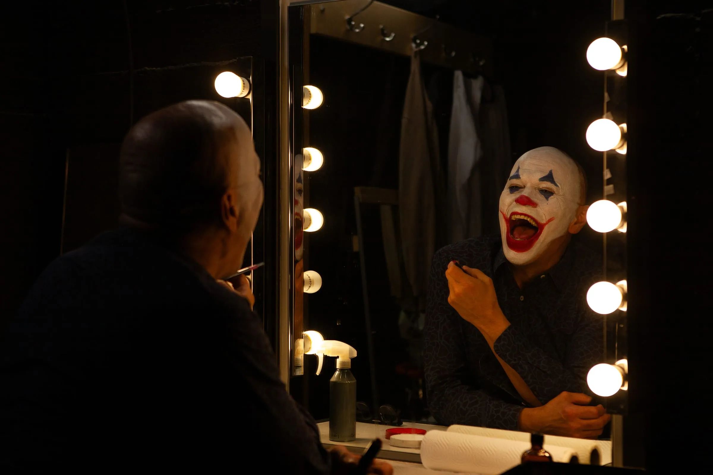 Backstage vor dem beleuchteten Schminkspiegel: Der Joker lacht, im Vordergrund eine zweite Person.