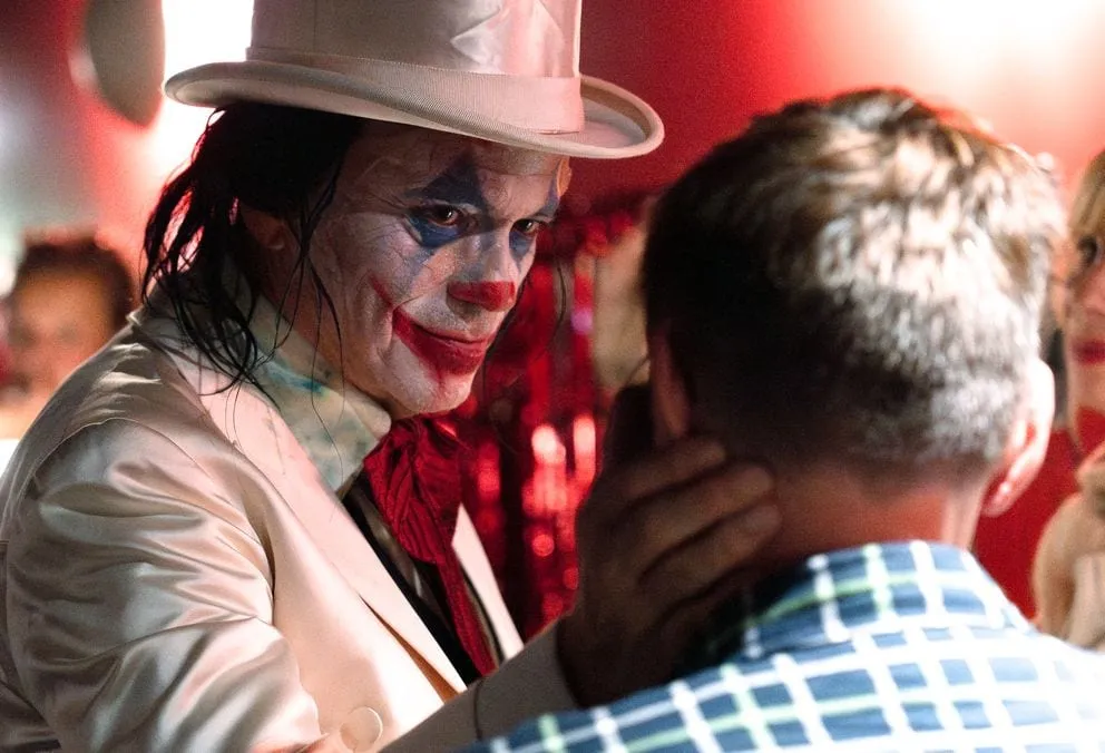 Der Joker mit weißem Zylinder beugt sich im roten Licht zu einem Zuschauer hinunter.