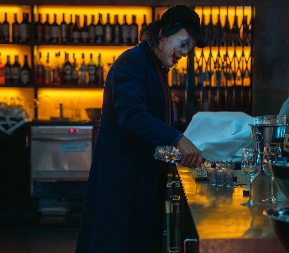 Der Joker steht hinter der Bar und schenkt ein, dahinter ein warm beleuchtetes Flaschenregal.