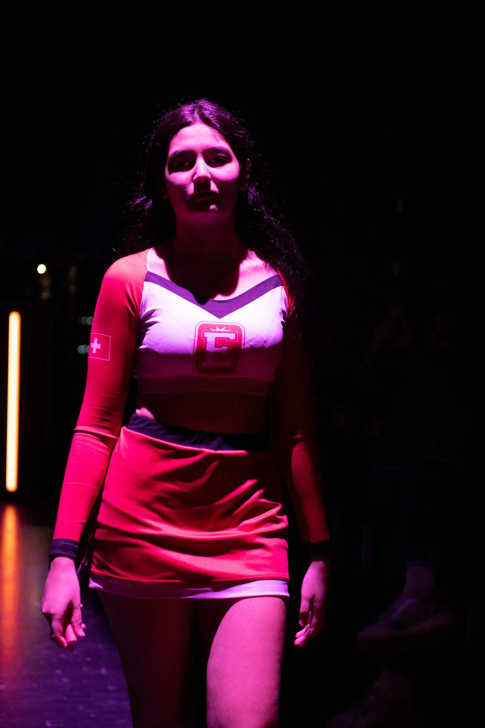Eine Darstellerin in rosa Top und Rock steht in magentafarbenem Licht auf der Bühne.