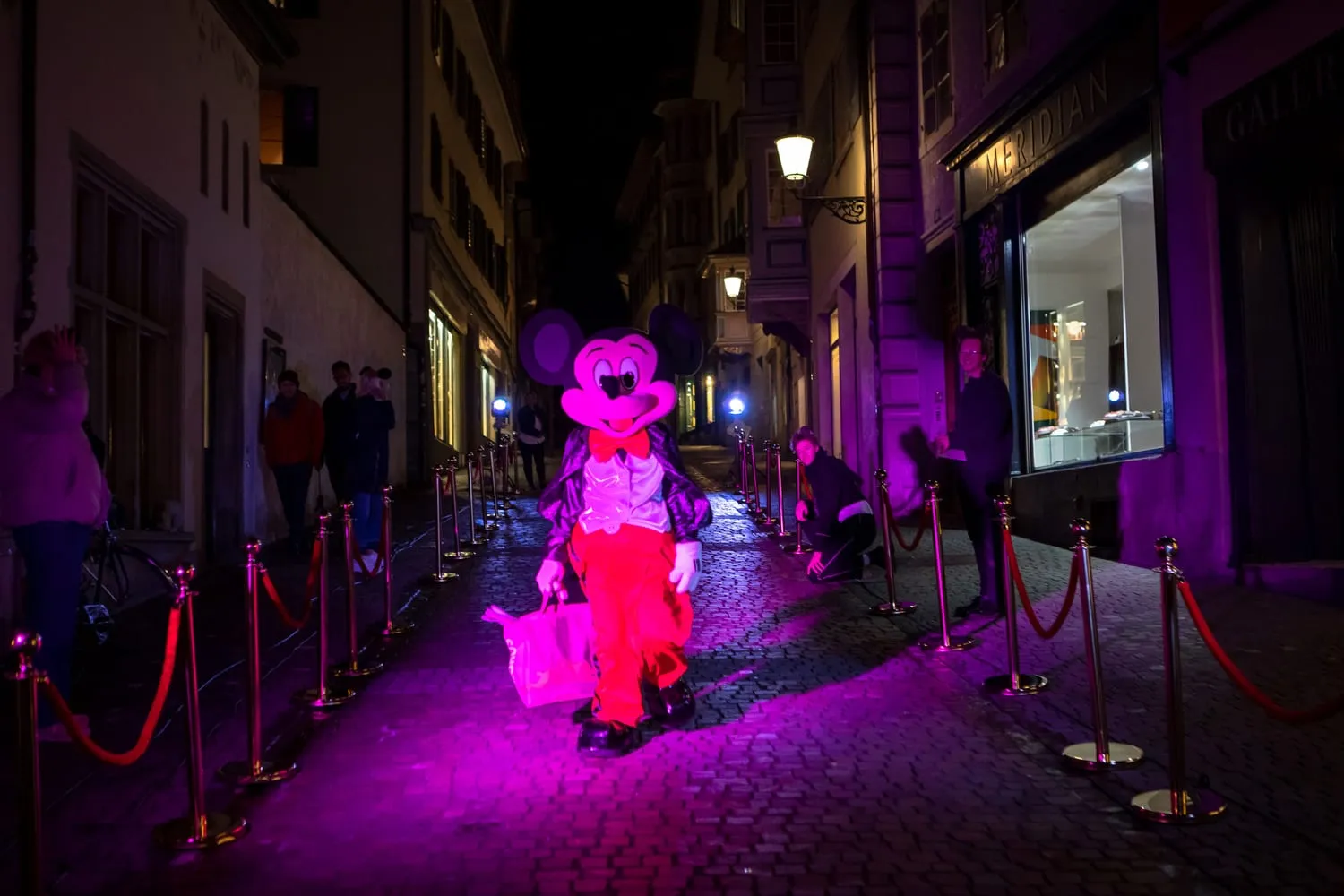 Eine Figur im Minnie-Maus-Kostüm geht magentafarben angestrahlt durch die nächtliche Gasse.