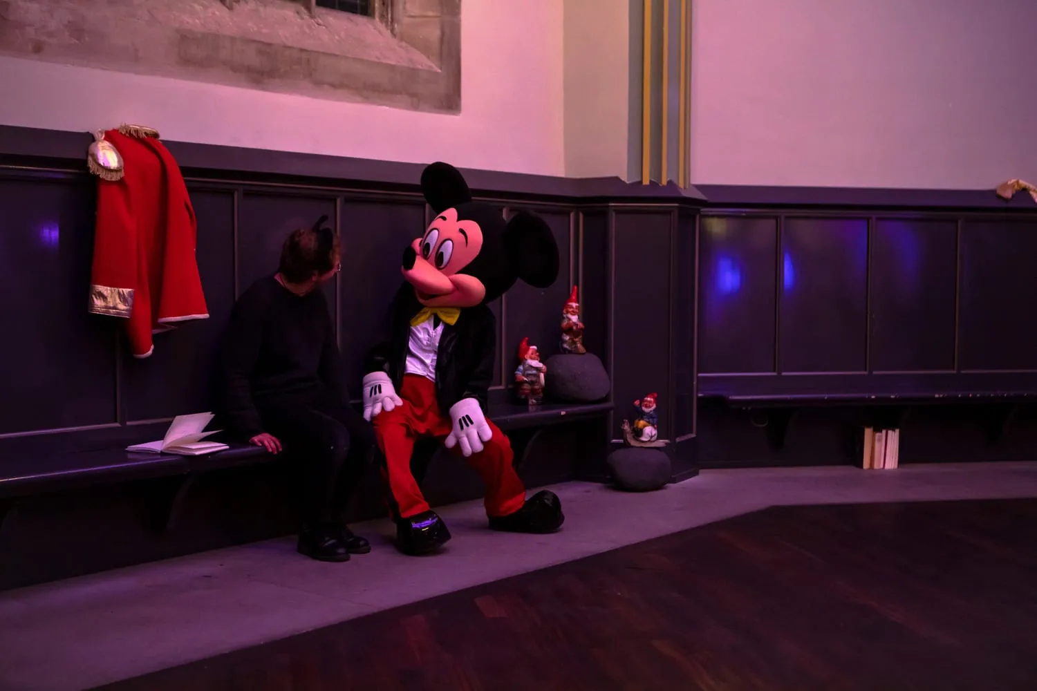 Zwei Figuren im Micky-Maus-Kostüm vor einer holzvertäfelten Wand, daneben hängt ein roter Mantel.