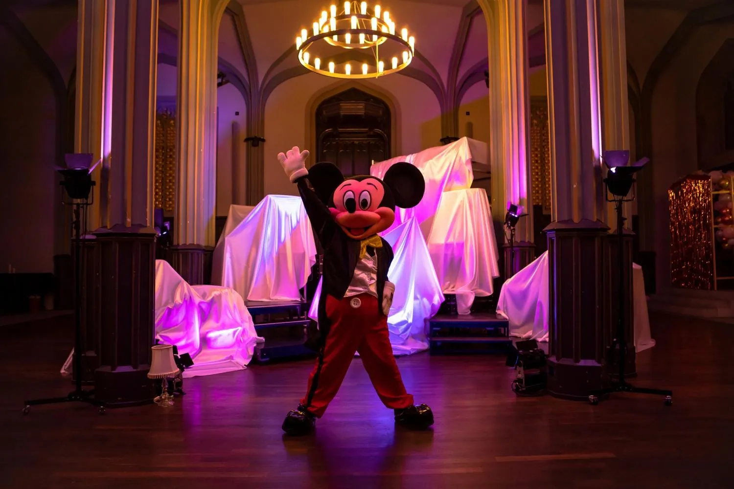Eine Figur im Micky-Maus-Kostüm hebt den Arm im Saal unter einem Kronleuchter, ringsum weiß verhüllte Objekte in violettem Licht.