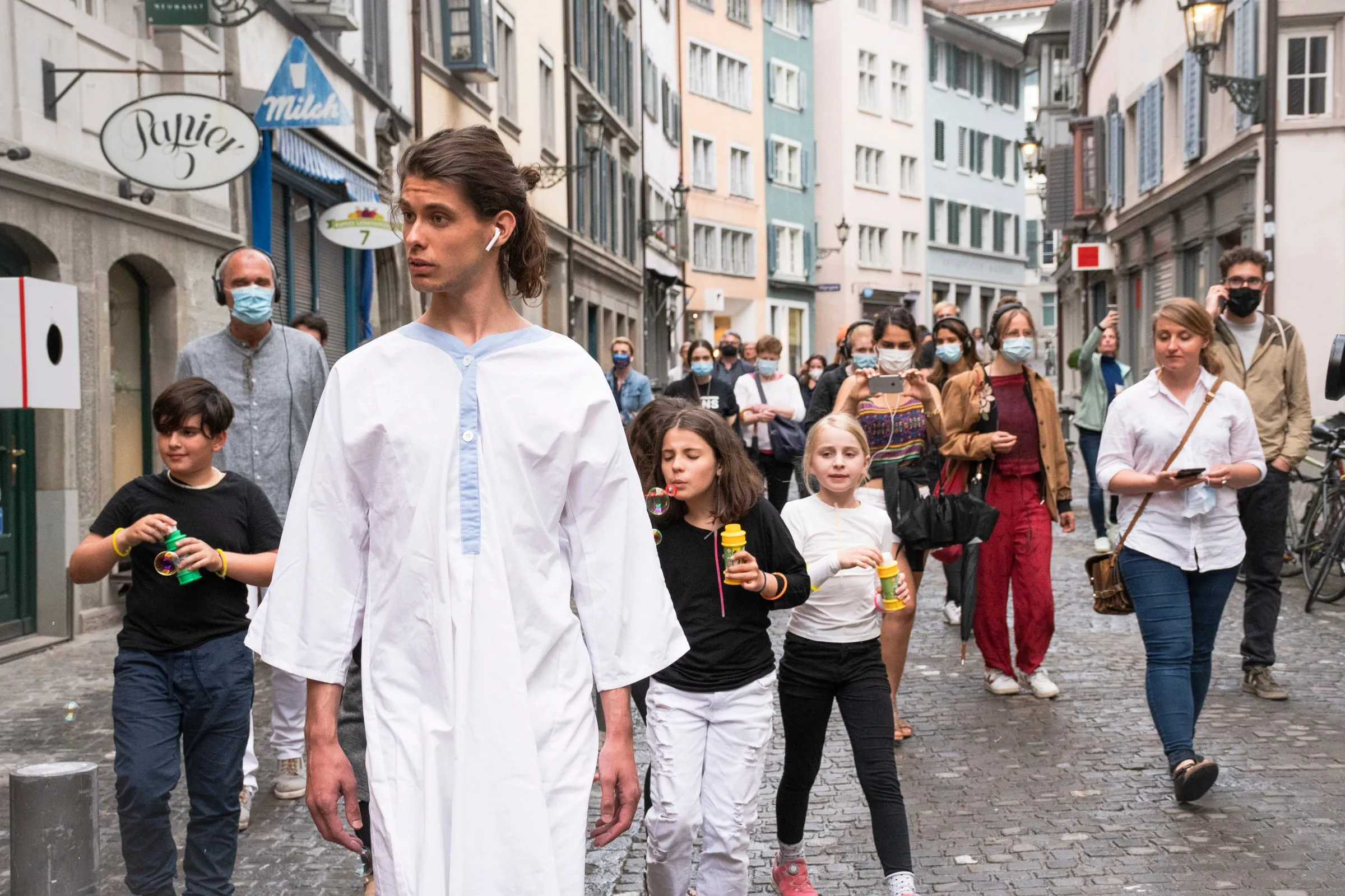 Eine Person in weißem Gewand führt eine Prozession durch eine Gasse in der Zürcher Altstadt, Passanten sehen zu.